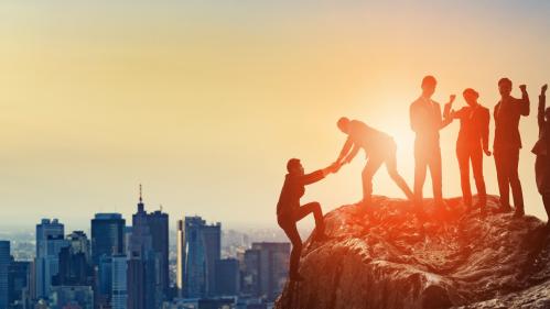 Cityscape in background with people at the top of a rock, one helping another climb to the top