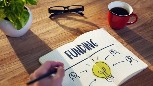 Desk with coffee mug, eyeglasses and open notebook with Funding written above an illustration of a light bulb